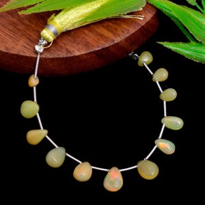 Puede incluir: Un collar de cuentas de ópalo en forma de pera, verde claro y naranja, ensartadas en un alambre de plata. Una borla amarilla con detalles plateados está unida en la parte superior. Las cuentas se muestran sobre un fondo oscuro.