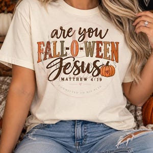 Peut inclure: Un t-shirt à manches courtes de couleur crème avec une impression graphique portant le texte "are you FALL-O-WEEN Jesus" dans des tons marron et orange. Le motif inclut une petite illustration de citrouille orange et le texte "MATTHEW 4:19".
