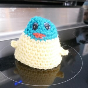 May include: A handmade crochet plush toy in the shape of a small creature. The top is turquoise, the body is cream, and it has black eyes and a red mouth. The toy has small arms and is sitting on a black surface.