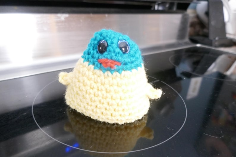 May include: A handmade crochet plush toy in the shape of a small creature. The top is turquoise, the body is cream, and it has black eyes and a red mouth. The toy has small arms and is sitting on a black surface.