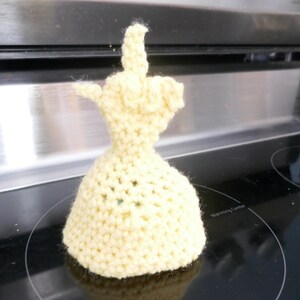 May include: A handmade, light yellow crocheted sculpture of a hand giving the middle finger. The sculpture is sitting on a black stovetop. The crocheted piece has a flared base and detailed fingers.