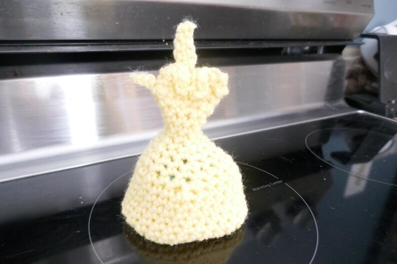 May include: A handmade, light yellow crocheted sculpture of a hand giving the middle finger. The sculpture is sitting on a black stovetop. The crocheted piece has a flared base and detailed fingers.