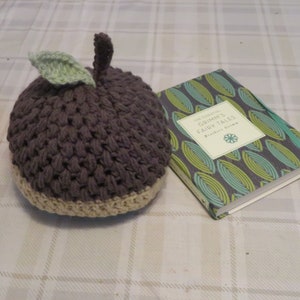 May include: A crocheted brown and beige apple hat with a green leaf on top. The hat is sitting on a white and gray plaid tablecloth next to a book titled "The Essential Grimm's Fairy Tales" by Brothers Grimm.