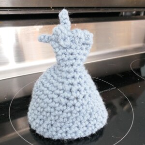 May include: A light blue crocheted oven mitt shaped like a hand giving the finger. The mitt is made of a soft, textured yarn and sits on a black stovetop. The mitt is a unique and humorous kitchen accessory.