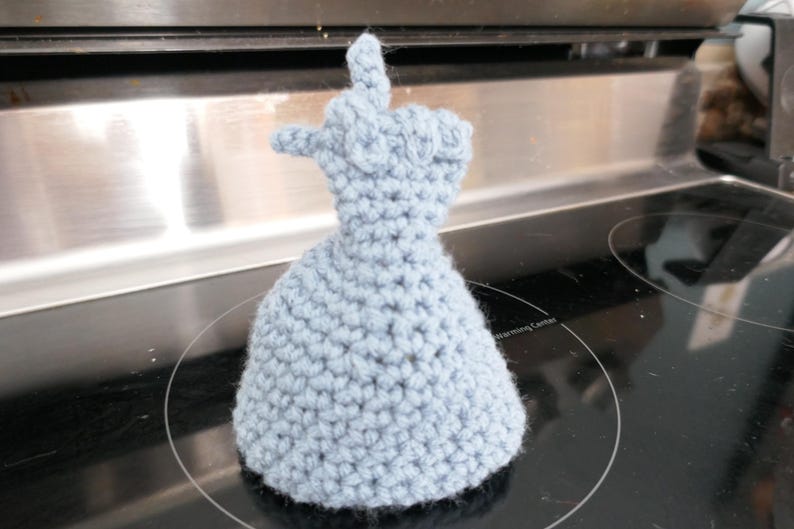 May include: A light blue crocheted oven mitt shaped like a hand giving the finger. The mitt is made of a soft, textured yarn and sits on a black stovetop. The mitt is a unique and humorous kitchen accessory.