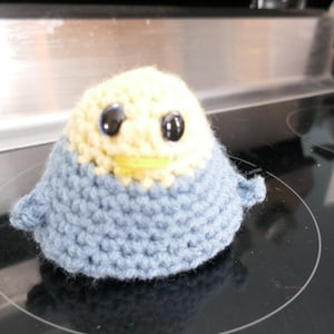 May include: A handmade crochet bird with a light blue body and a cream-colored head. The bird has large black eyes and a yellow beak. The craft project is sitting on a black stovetop.