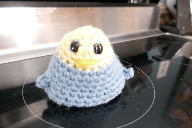 May include: A handmade crochet bird with a light blue body and a cream-colored head. The bird has large black eyes and a yellow beak. The craft project is sitting on a black stovetop.