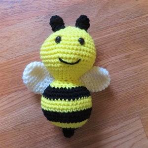 May include: A crocheted bee toy with black and yellow stripes, white wings, and black eyes. The bee has a smiling face.