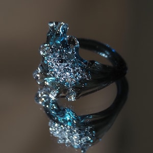 May include: A dark blue ring with an irregular, molten-like texture featuring a cluster of small, sparkling clear stones embedded in the centre. The surface has a glossy, reflective finish that catches the light, creating a deep, oceanic appearance.