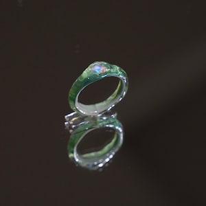 May include: A ring featuring a translucent green band with an irregular, organic texture. A small, round, iridescent stone with flashes of blue, orange, and pink is set into the top of the band. The ring has a smooth, reflective inner surface.