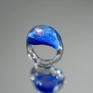 May include: A clear glass ring featuring a rounded top section infused with deep blue swirls and a small, iridescent, multi-colored fleck embedded within the glass. The band is transparent and smooth, creating a continuous, fluid, and organic shape.