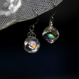 Può includere: Un paio di orecchini pendenti con perle di vetro trasparenti e arrotondate sospese a ganci in metallo color argento. Ogni perla di vetro contiene un piccolo inserto iridescente e multicolore che riflette la luce con vivaci tonalità di blu, verde e arancione.