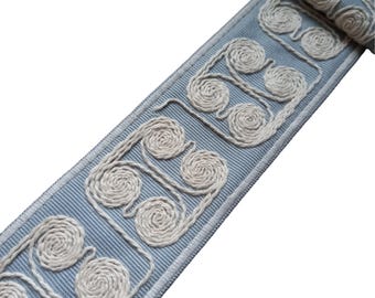 May include: A light blue trim with a white embroidered design of repeating swirls. The trim is woven and has a subtle texture.