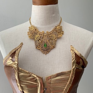 May include: A brown and gold corset with a gold filigree necklace featuring a green gemstone.
