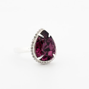 Peut inclure: Une bague en argent ornée d'une grosse pierre précieuse violet foncé en forme de poire. La pierre est entourée d'un halo de petites pierres claires. L'anneau de la bague est en argent poli.