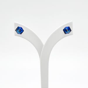 Peut inclure: Une paire de boucles d'oreilles clous cubiques bleues. Chaque boucle d'oreille présente un cristal bleu profond à facettes, monté sur un présentoir blanc. Les boucles d'oreilles ont un design géométrique et reflètent la lumière.
