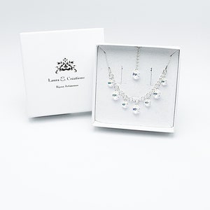 Puede incluir: Un conjunto de collar y pendientes de plata con piedras de cristal transparentes. Las joyas est&aacute;n empaquetadas en una caja blanca con el texto "Laura S. Creations Bijoux Artisanais".