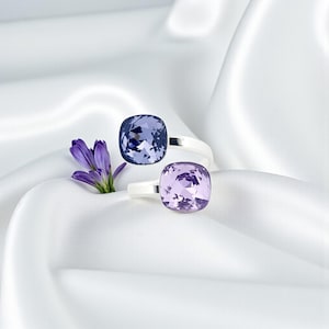 May include: Two silver rings, each with a large, square, faceted gemstone. One ring has a deep blue gem, the other a light purple gem. They are displayed on white, silky fabric, with a sprig of purple flowers.