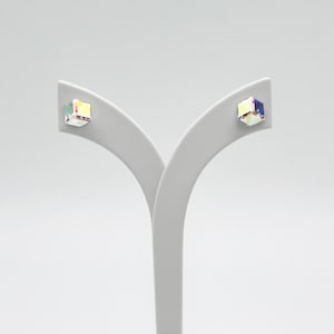Könnte beinhalten: Ein Paar schillernde, würfelförmige Ohrstecker. Jeder Ohrring ist ein kleines, quadratisches Prisma mit einer schimmernden, mehrfarbigen Oberfläche. Die Ohrringe werden auf einem weißen, gebogenen Schmuckständer vor einem schlichten weißen Hintergrund präsentiert.