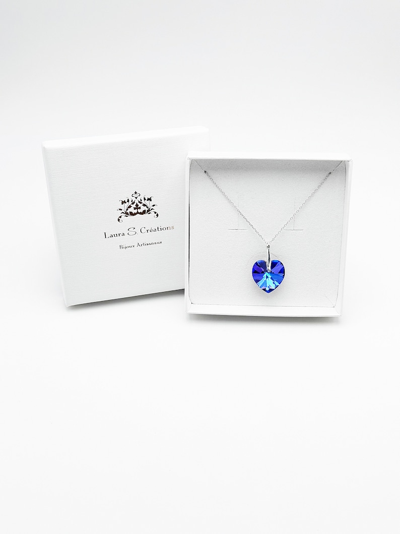 May include: A silver necklace with a blue crystal heart pendant in a white box. The box has the text "Laura & Cr&eacute;ations Bijoux Artisanaux" printed on it.