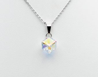 Pode incluir: Um colar de prata com um pingente cúbico. O pingente é um cristal transparente com tons iridescentes de arco-íris. A corrente delicada pende de uma pequena argola de prata.