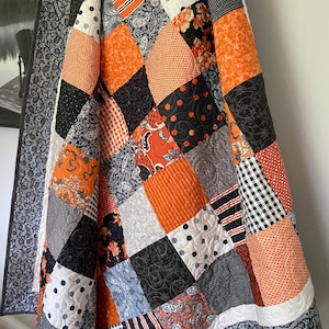 May include: A patchwork quilt featuring a variety of square fabric blocks in orange, black, white, and gray. The patterns include polka dots, stripes, gingham, paisley, and floral motifs, all joined with visible machine quilting in a swirling pattern.