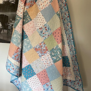 May include: A patchwork quilt with a variety of floral and geometric patterns in shades of pink, blue, green, and peach. The quilt is made of squares and has a blue border. The quilt is hanging on a wall.