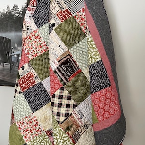 May include: A patchwork quilt with a variety of fabrics in red, green, black, and white. The quilt features a variety of patterns, including plaid, polka dots, and floral prints. The quilt is made with a variety of fabrics, including cotton and flannel. The quilt is edged with a gray fabric.