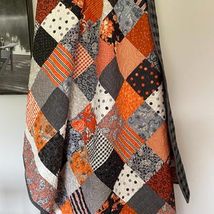 May include: A patchwork quilt featuring a variety of square fabric blocks in orange, black, white, and gray. The patterns include floral prints, polka dots, gingham, stripes, and paisley designs, all stitched together with a dark gray plaid border.