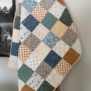 May include: A patchwork quilt with a variety of square patterns in shades of blue, green, tan, and brown. Some squares feature letters, stars, and other designs. The quilt is draped over a wooden ladder.