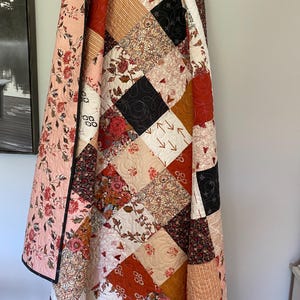 May include: A patchwork quilt featuring a variety of square fabric blocks in shades of rust, black, cream, and soft pink. The quilt displays diverse patterns including floral prints, geometric lines, and small heart motifs, all finished with intricate quilting.