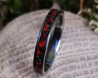 Puede incluir: Un anillo negro con una fina banda roja de purpurina incrustada. El anillo está sobre una superficie de madera.