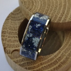 May include: A silver ring with a blue and white glitter inlay. The ring is sitting in a wooden ring box.