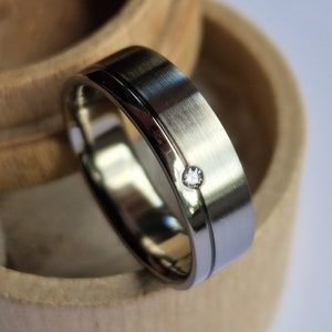Op de afbeelding: Een zilveren trouwring met een enkele diamant in het midden. De ring ligt in een houten ringendoos.