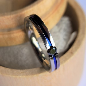 May include: A silver ring with blue and black stripes and a black gemstone in the centre. The ring is sitting in a wooden box.