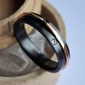 Puede incluir: Una banda de boda negra y oro rosa con un solo diamante engastado en el centro. La banda está apoyada en una caja de madera.