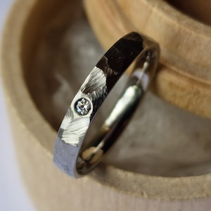 Puede incluir: Una banda de plata con un pequeño diamante engastado en el centro, descansando en una caja de anillo de madera.