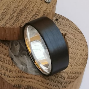 May include: A black and silver ring in a wooden ring box. The ring is made of carbon fiber and has a silver band.