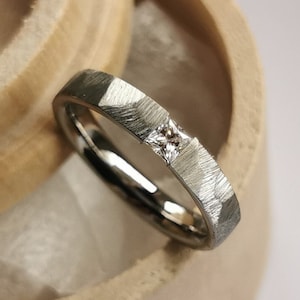 Op de afbeelding: Een zilveren band met een gehammerde textuur en een kleine diamant in het midden. De ring zit in een houten doos.