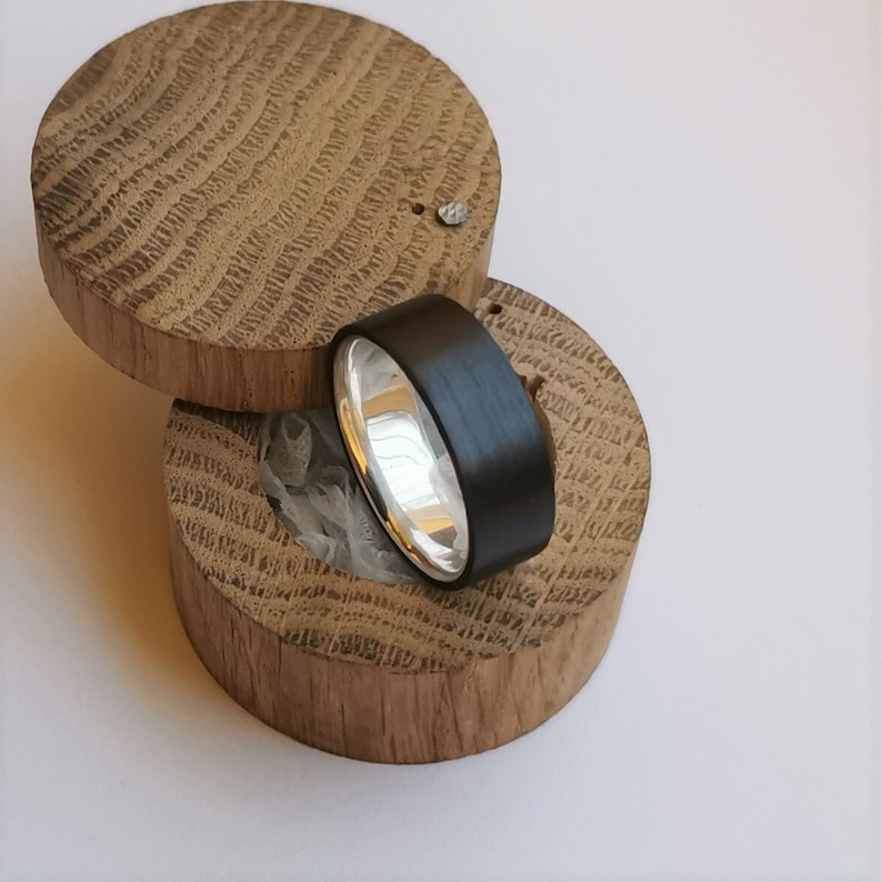 May include: A black and silver ring in a wooden ring box. The ring is made of carbon fiber and has a silver band.