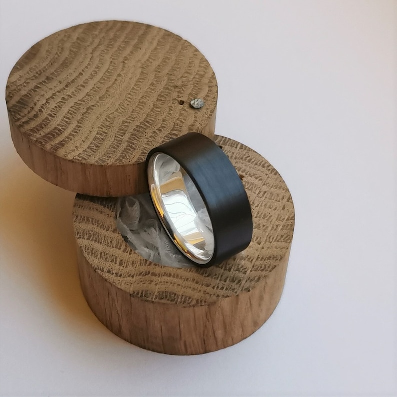 May include: A black and silver ring in a wooden box. The ring is made of a black material with a silver band. The box is made of wood with a natural finish.
