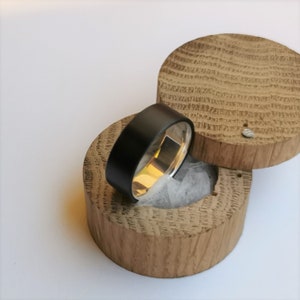 May include: A black and silver ring in a wooden ring box. The ring is made of a black material with a silver band.