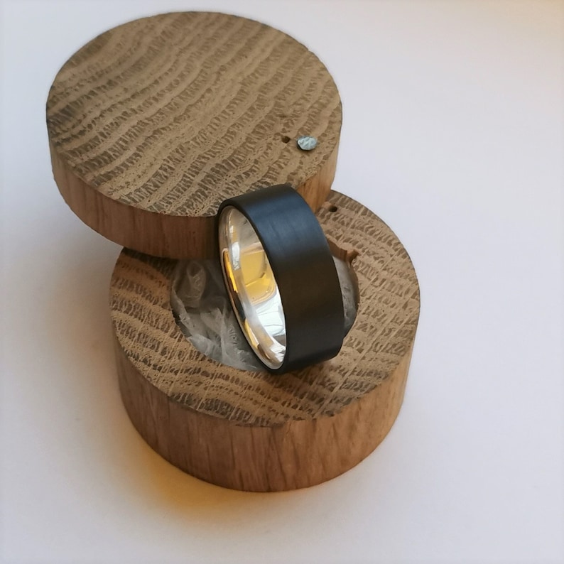 May include: A black and silver ring in a wooden ring box. The ring is made of a black metal with a silver band.