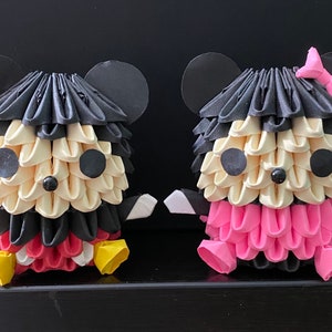 May include: Two origami Mickey Mouse figures, one in black and red and the other in pink and black. The figures are sitting on a black surface.