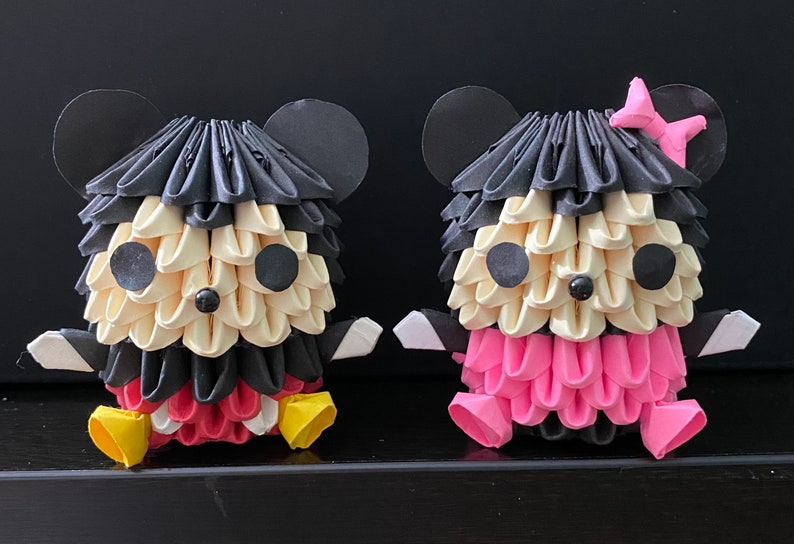 May include: Two origami Mickey Mouse figures, one in black and red and the other in pink and black. The figures are sitting on a black surface.