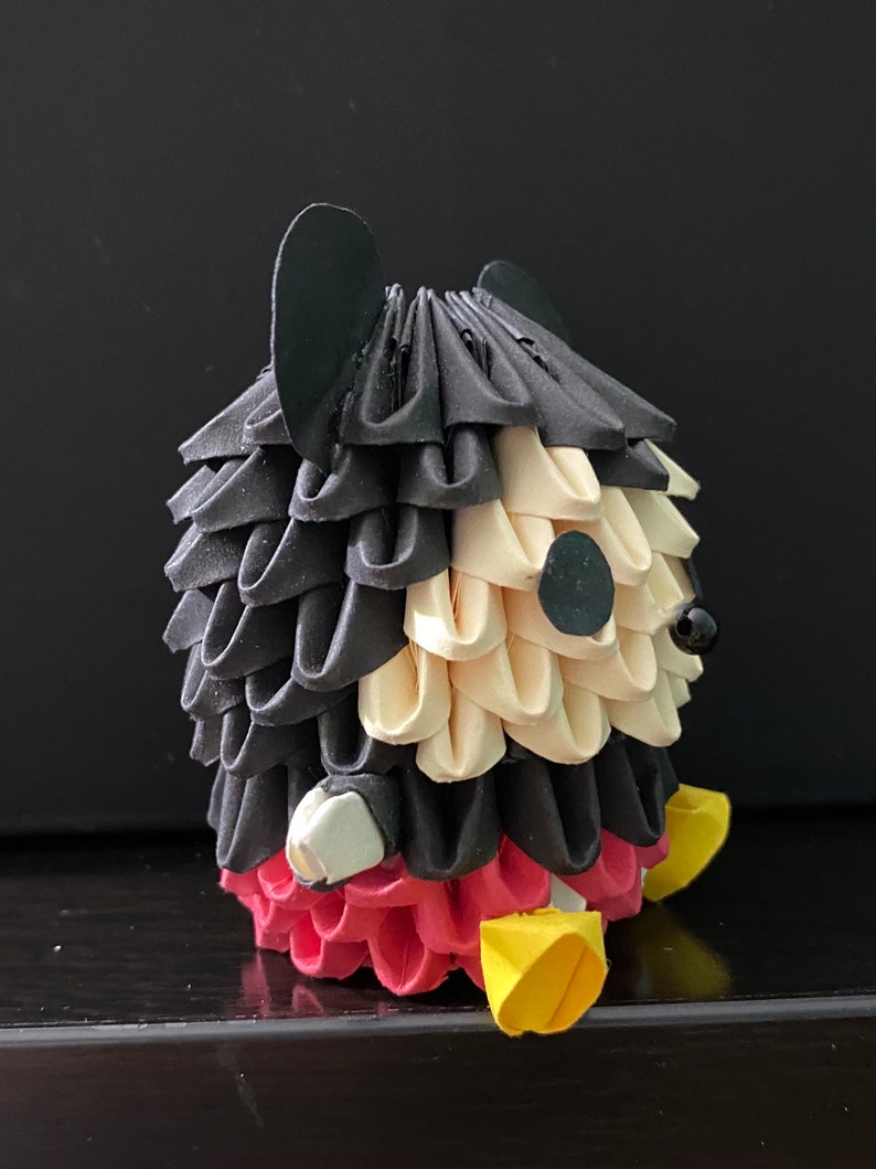 May include: An origami Mickey Mouse figure made from black, white and red paper. The figure is folded into a three-dimensional shape.