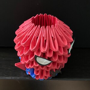 Może przedstawiać: Czerwona rzeźba origami głowy Spider-Mana. Rzeźba wykonana jest ze złożonego papieru i ma białe oko oraz czarny wz&oacute;r pajęczyny.