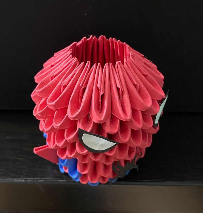 Może przedstawiać: Czerwona rzeźba origami głowy Spider-Mana. Rzeźba wykonana jest ze złożonego papieru i ma białe oko oraz czarny wz&oacute;r pajęczyny.