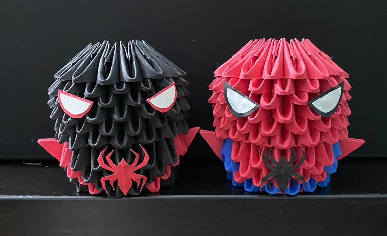 Może przedstawiać: Dwie figurki origami Spider-Mana, jedna w kolorze czarnym i czerwonym, a druga w czerwonym i niebieskim. Obie figurki mają białe oczy i czarny symbol pająka na klatce piersiowej.