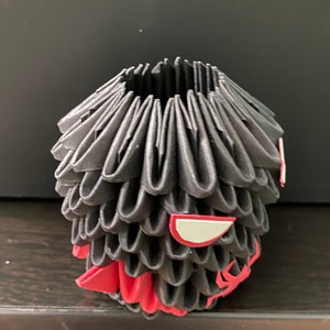 Może przedstawiać: Czarna rzeźba origami z czerwonymi i białymi akcentami. Rzeźba ma kształt głowy ze stylizowaną twarzą.
